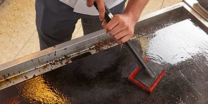 chef scouring and scrubbing a griddle with a pad and holder tool
