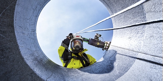 Worker in protective gear looking down into a confined space entry portal.
