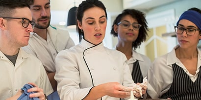 Four onlooking commercial kitchen workers surround a chef demonstrating a food preparation technique.
