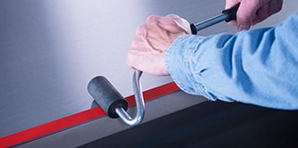 A worker uses a J-Roller to apply pressure to a tape on a metal bar
