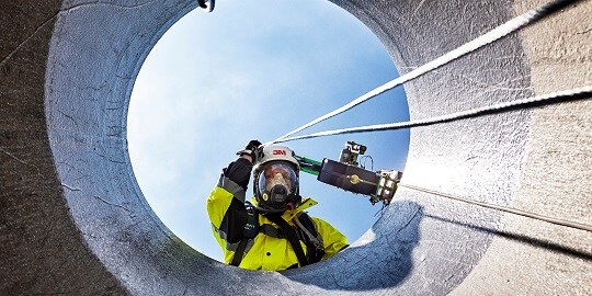 Worker in protective gear looking down into a confined space entry portal.
