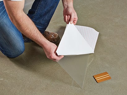 A man kneeling on the floor peels 3M™ Scotchgard™ Surface Protection Film from its backing.

