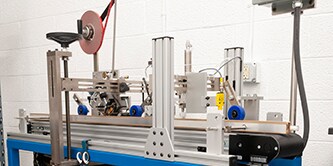 Image of a complex machine for applying tape to parts as they pass underneath on a conveyor
